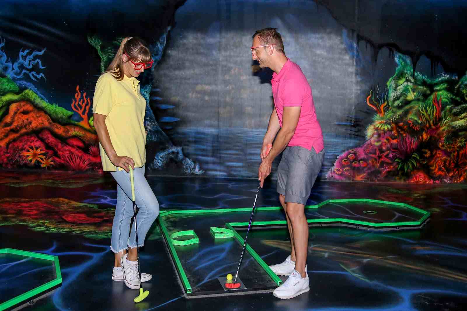 Eine Frau und ein Mann mit 3D-Brillen spielen Minigolf vor einer leuchtenden Unterwasser-Wandmalerei
